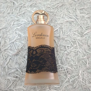 Loveliness Sensuelle parfymflaska - Parfymflaska i frostat glas med guldig kork och elegant svart spetsdetalj runt flaskan. Flaskan har en mjukt böjd form och texten 'Loveliness Sensuelle' på framsidan. Perfekt för dig som gillar lyxiga och feminina dofter.
