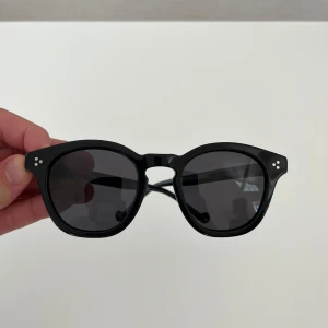 Svarta runda solglasögon - Snygga svarta solglasögon med rundad form och mörka glas. Bågarna är i blank plast och har små silvriga detaljer vid hörnen. Box till glasögon ingår.