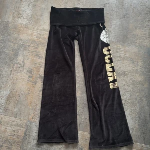Svarta foldover velourbyxor från PINK - Svarta mjukisbyxor i velour från PINK med bred midja och glittriga detaljer. Ena benet har stora guldiga bokstäver och ett mönstrat hundmotiv i strass. Byxorna har bootcut-modell och är riktigt sköna och snygga för en chill look. Byxorna är lågmidjade och när man foldover bandet syns det en Victorias Secret hund.