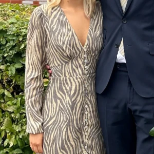 Zebramönstrad klänning från Adoore - Snygg zebramönstrad klänning från Adoore i beige och brunt. Klänningen har v-ringning, långa ärmar och knappar framtill. Den är figurnära upptill och har en lätt utställd kjol som slutar under knät. Perfekt för dig som vill sticka ut med ett coolt mönster. Använd en gång. Nypris 1700kr. 