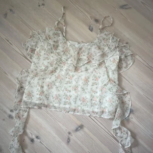 Blommig volangtopp från Gina Tricot - Supersöt blommig topp med volanger och tunna axelband från Gina Tricot. Topp i ljus, nästan vit färg med små rosa och gröna blommor. Luftigt material, Perfekt för sommarens alla tillfällen.