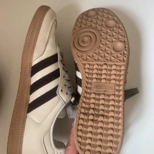 Adidas Samba sneakers i vitt skinn - Adidas Samba sneakers i vitt skinn med bruna ränder och klassisk gummisula. Skorna har breda, brunvita snören och gulddetalj med Samba-tryck på sidan. Perfekt retrostil med rund tå och låg profil.