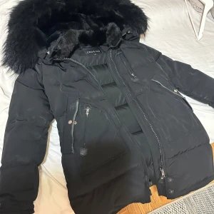 Svart dunjacka med päls från Cedrico - Säljer en svart dunjacka från Cedrico med stor fluffig päls på huvan. Jackan har flera dragkedjor, stora fickor och en quiltad design. Perfekt för kalla dagar och riktigt snygg streetstil. .Det finns en liten defekt som visas men den sitter inuti jackan. Varm jacka.