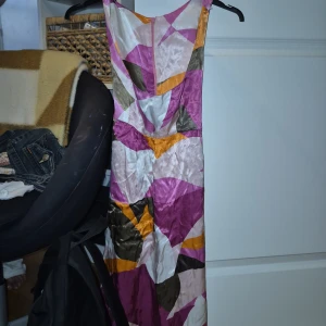 Färgglad långklänning från Bershka - Säljer en maxad långklänning från Bershka med coolt geometriskt mönster i lila, rosa, orange, beige och olivgrönt. Klänningen har omlottknytning i midjan och är ärmlös. Materialet är glansigt och känns lyxigt, perfekt för dig som vill sticka ut.
