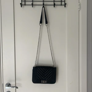 Svart quiltad axelväska med kedja - Snygg svart axelväska med quiltad design och silverfärgad kedja som axelrem. Väskan har ett stilrent rektangulärt spänne i metall och ett innerfack med dragkedja.