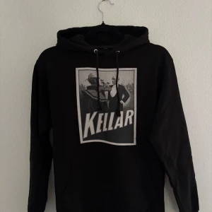 Svart hoodie ,Kellar, The Cool Elephant - Svart hoodie med stor Kellar-print på bröstet och snörning i huvan. På ryggen finns ett litet vitt tryck med The Cool Elephant-loggan. Tröjan har lång ärm och klassisk känguruficka framtill. Perfekt för dig som gillar streetwear och coola tryck.