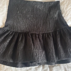 Svart glittrig volangkjol från Zara - Supersnygg svart kortkjol från Zara med glittrig finish och volang nertill. Kjolen har en crinklad struktur som ger en cool look och är perfekt för dig som vill sticka ut. Hög midja och figurnära passform upptill.