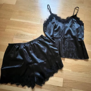 Svart sidenpyjamas med spets - Supersnygg svart pyjamas i glansigt sidenmaterial med tunna axelband och spetsdetaljer längs urringningen på linnet. Perfekt för dig som gillar en elegant och chic vibe. Aldrig använd!
