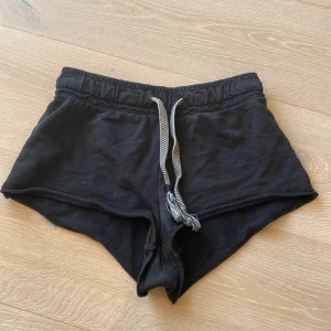 Svarta shorts med snörning från Bershka - Svarta mjuka shorts från Bershka i bomull med elastisk midja och vitmönstrad snörning framtill. Perfekta för sommardagar eller chill hemma. Klassisk sportig look med korta ben och relaxed fit.