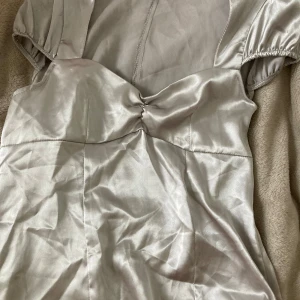 Silver topp Theos Hero - Säljer en snygg topp  från Theos Hero i Silver satin. Klänningen har korta puffärmar med resår, en lätt rynkad detalj vid bysten och en slät, figurnära siluett. Perfekt för dig som gillar stilren och glansig look.