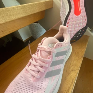 Adidas rosa sneakers med Boost-sula - Snygga rosa Adidas sneakers med mesh-ovandel och klassiska tre ränder i ljusgrått på sidorna. Skorna har vit Boost-mellansula för extra komfort och svart/rosa yttersula med Continental-gummi för bra grepp. Perfekta för dig som gillar sportig stil och vill sticka ut.