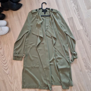 Tunn Olivgrön trenchcoat från Forever 21 - Snygg olivgrön trenchcoat från Forever 21 med långa ärmar, stora fickor och coola axelremmar. Jackan har bälte i ärmsluten och en avslappnad, loose passform. Perfekt för lager-på-lager och ger en chill men stilren vibe.