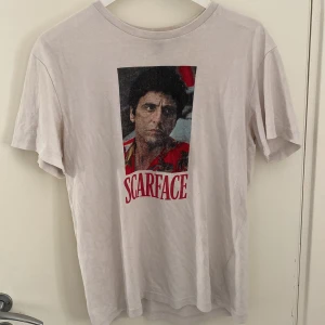 Scarface t-shirt med tryck - Vit t-shirt med Scarface-tryck på framsidan i rött och svart. Klassisk rund hals och korta ärmar. Materialet känns som mjuk bomull, perfekt för dig som gillar film och streetwear. Passformen är normal och t-shirten har en clean look baktill utan tryck.