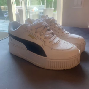 Vita sneakers från Puma med platåsula - Snygga vita Puma sneakers med svart sidodetalj och guldfärgad logga. Skorna har en platåsula och klassisk snörning. Perfekta för dig som vill ha en sportig och trendig look.