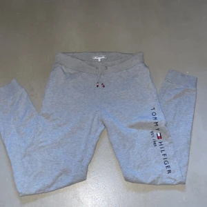Grå mjukisbyxor Tommy Hilfiger - Snygga grå mjukisbyxor från Tommy Hilfiger med logga och text längs benet. Byxorna har snörning i midjan, muddar vid bensluten och en bakficka med liten flagga. Perfekta för chill eller träning.