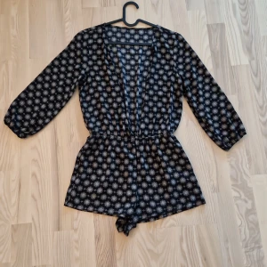 Svart mönstrad playsuit med shorts - Svart playsuit med shorts och långa ärmar, täckt av ett vitt geometriskt mönster. Resår i midjan för skön passform och v-ringad hals. Lätt och luftig, perfekt för varma dagar eller festival. Materialet känns som tunn bomull eller viskos.