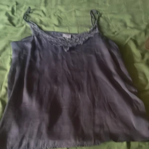 Grått  linnetopp med spetskant - Säljer en snygg mörklila linnetopp med tunna axelband och fin spetsdetalj längs urringningen. Toppen har en slät, glansig yta och är perfekt för dig som gillar en enkel men ändå elegant stil.