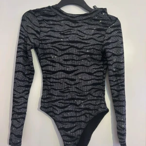 Glittrig zebramönstrad långärmad body - Säljer en svart och silver body med glittrigt zebramönster. Toppen har lång ärm och en djup v-ringad rygg som ger en snygg och edgy look. Materialet är stretchigt och skönt, perfekt för att sticka ut. Passformen är tight och figurnära.