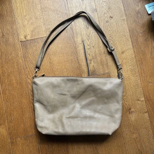 Beige axelväska i skinn - Stilren beige axelväska i mjukt skinn med justerbar rem och dragkedja upptill. Insidan är fodrad i vinrött tyg och har en praktisk innerficka. Perfekt storlek för det viktigaste och enkel att matcha till olika outfits.