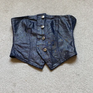 Svart glittrig jeanskorsett med knappar - Snygg svart glitter  korsett i jeansmaterial med metallknappar framtill. Modellen har en cool, spetsig nederkant och markerade sömmar som ger en edgy vibe. Perfekt att styla över en skjorta eller t-shirt för en trendig look eller ha den som den är!!
