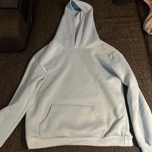 Ljusblå hoodie med butterfly-tryck - Säljer en ljusblå hoodie med stor fjäril och texten 'BUTTERFLY' på ryggen. Hoodien har en klassisk känguruficka framtill och mjukt material, perfekt för chill dagar. Långärmad och med huva för extra mysfaktor. Snyggt tryck som sticker ut och ger street vibe.