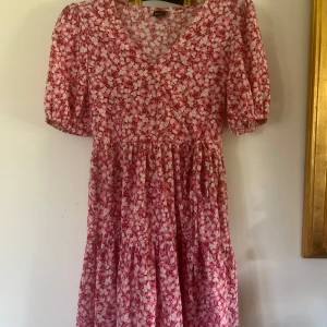 Rosa blommig omlottklänning Gina Tricot - Supersöt kort omlottklänning från Gina Tricot i rosa med vitt blommigt mönster. Klänningen har puffiga korta ärmar, v-ringning och knyt i midjan. Materialet är mjukt och lätt, Passformen är normal.