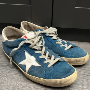 Blå Golden Goose Superstar sneakers - Golden Goose Superstar sneakers i blå mocka med vit stjärndetalj på sidan, vita snören och vit häl. Klassisk retrostil med platt sula och rund tå. Snygg kontrast mellan blå mocka och vita detaljer, perfekt för dig som gillar streetstyle.