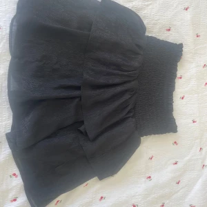 Svart volangkjol med smock - Supersöt svart kortkjol med bred smock i midjan och dubbla volanger som ger extra fluff. Kjolen har en lätt glansig yta och är perfekt för dig som gillar att sticka ut med en cool och trendig look. Passar dig som vill ha något bekvämt men ändå snyggt.