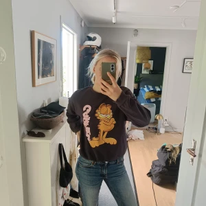 Mörkgrå Garfield sweatshirt - Snygg mörkgrå sweatshirt med stort tryck av Garfield och rosa text på framsidan. Rund hals, ribbade muddar och långärmad. Mjuk och skön bomullsmix som passar till jeans eller chill-outfit.