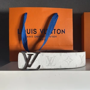 Vit Louis Vuitton skinnbälte - Säljer ett stilrent vitt Louis Vuitton bälte i skinn med klassisk LV-logga i silver på spännet. Bältet har diskret monogrammönster längs hela remmen och en elegant, rak form. Perfekt för dig som vill ha en lyxig accessoar med ikonisk design.