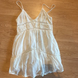 Vit omlottklänning från Zara - Supersöt vit kortklänning från Zara med tunna axelband och omlott-design. Klänningen har fina spetsdetaljer och ett lätt, luftigt tyg som ger en bohemisk vibe. Perfekt för varma dagar och sommarhäng. Säljes i storlek S.