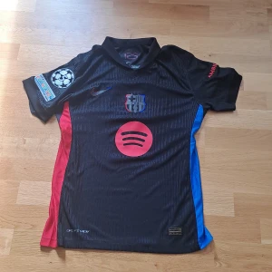 FC Barcelona Lamine Yamal tröja Nike player version  - Säljer en svart FC Barcelona fotbollströja från Nike med Lamine Yamal och nummer 19 på ryggen. Tröjan har röda och blå detaljer på sidorna, klubbmärke på bröstet, Spotify-logga och Champions League-märke på ärmen. Tillverkad i lätt och ventilerande material. 