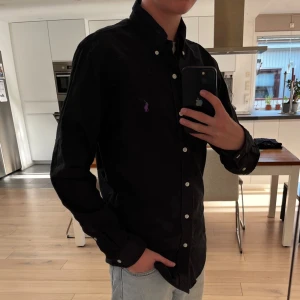 Svart skjorta från Polo Ralph Lauren - Klassisk svart skjorta från Polo Ralph Lauren med smal passform. Skjortan har långa ärmar, vita knappar och en broderad lila logga på bröstet. Tillverkad i bomull och passar perfekt till dig som gillar stilrena plagg.