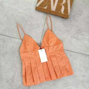 Randig orange festtopp från Zara - Superfin orange linnetopp från Zara med smala axelband och vita ränder. Toppen har v-ringad skärning, veckad nederdel och dragkedja i sidan. Perfekt för dig som gillar en fräsch och somrig vibe.