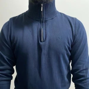 Marinblå stickad polotröja Holebrook - Snygg marinblå stickad polotröja från Holebrook Sweden med ribbad hög krage och half zip-dragkedja. Tröjan har diskret broderad logga på bröstet och är långärmad. Perfekt för dig som gillar stilren och klassisk design.