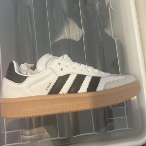 Adidas Samba OG sneakers vit/svart - Klassiska Adidas Samba OG sneakers i vitt skinn med svarta ränder och detaljer. Snygg gummisula i beige, vit snörning och rund tå. Tidlös streetstil med retrovibbar, perfekt till jeans eller shorts. Ikonisk logga på plösen och sidan.