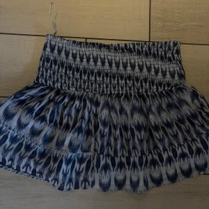 Blåvit kortkjol med mönster - Säljer en blåvit kortkjol med elastisk midja och ett coolt abstrakt mönster. Kjolen har volangkant och är gjord i ett lätt och luftigt material som passar perfekt för varma dagar. Supersnygg att matcha med en enkel topp och sneakers.