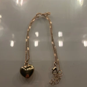 Guldigt halsband med hjärthänge - Snyggt halsband i guldton med ett blankt hjärthänge. Kedjan har små detaljer och justerbar längd med flera ringar vid låset. Perfekt accessoar för att lyfta din outfit med en söt och romantisk touch.