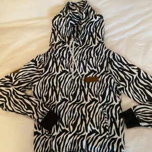 Zebramönstrad hoodie Fallen Star XS - Snygg hoodie från Fallen Star London i zebramönster, svart och vit. Jackan har huva med snörning, ribbade muddar och dragkedja i fickan. 