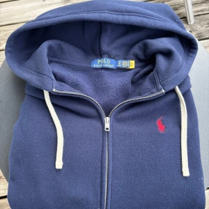 Ralph Lauren zip hoodie marinblå med kvitto! - Snygg marinblå hoodie från Ralph Lauren, köpt på Zalando skriv till mig så får du kvitto. Klassisk röd broderad logga på bröstet. Tröjan har huva med vita snören och dragkedja framtill. Mjuk insida i fleece och normal passform, perfekt för chill eller när du vill ha en clean look.