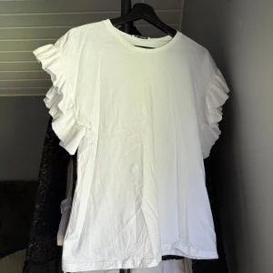 Vit topp med volangärmar från Zara - Säljer en vit topp från Zara i storlek S med snygga volangdetaljer på ärmarna. Modellen är rak och har rund halsringning. Perfekt för dig som gillar en clean look med lite extra edge på axlarna. Materialet känns mjukt och skönt mot huden.🩷