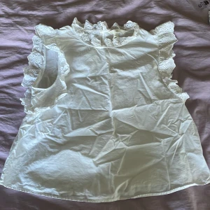 Vit blus med volang från H&M - Supersöt vit blus från H&M i bomull med volangdetaljer och broderad spets runt ärmhål och hals. Ärmlös modell med romantisk känsla och rund halsringning. Perfekt för dig som gillar en feminin och trendig look.