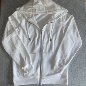 Vit hoodie med dragkedja och huva - Säljer en fräsch vit hoodie med dragkedja och huva. Tröjan har långa ärmar, snörning vid huvan och två fickor framtill. Perfekt för chill dagar eller när du vill ha en enkel och clean look. Materialet känns mjukt och bekvämt mot huden. Tråden ingår ej!