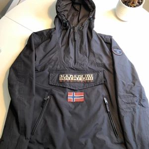 Pullover-jacka från Napapijri - Svart supersnygg pullover jacka från Napapijri i storlek S. Jackan är rätt stor i storleken så du som vanligtvis använder Medium kan absolut använda denna. Jackan går att spänna åt i midjan och luvan för anpassad passform vilket gör att den kommer sitta perfekt för just dig 🔥 Tål alla typer av klimat och har rymliga och bra fickor