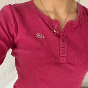 En tröja i pop-rosa färg med ett silvrigt snirkligt broderat B på bröstet. Knappar som går att knäppa eller ha lösa. Använd men bra skick. 