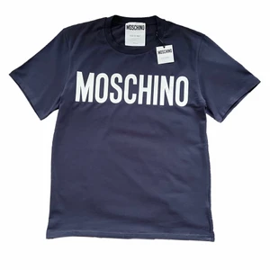 Moschino T-shirt - En t-shirt från Moschino i storlek M som aldrig är använd. Jag köpte den från Johnells, de i kassan sa att någon förut hade försökt snatta denna och därför har den små hål där larmet satt i nedre hörnet som ni ser på bilden. Detta är dock inget som syns och ifall man har den instoppad är det omöjligt att se det. Skriv privat för fler bilder eller för bilder på hur den sitter på mig. Jag kan mötas upp i Stockholm eller skicka den om köparen står för frakt.