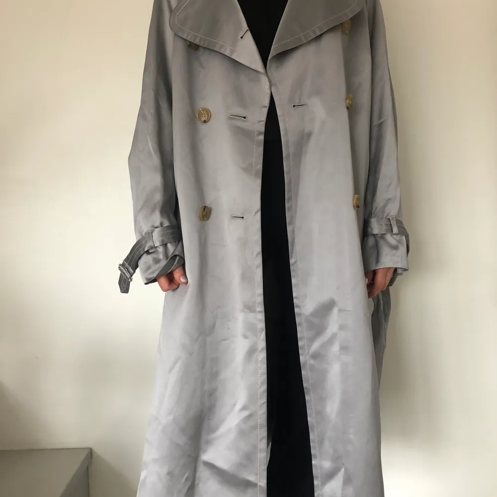 Grå/silvrig trenchcoat från Tiger of Sweden i modellen Zparra, storlek 38. Tunn och härlig till våren eller svala sommarkvällar men funkar även sen höst med stickat under. Liten fläck från handdesinfektion som inte syns när man har på sig den. Nypris var 5000kr.. Jackor.