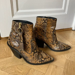 ormskinn cowboy boots stl 36 - Boots i ormskinnsimitation från & Other Stories i stl 36. I använt skick men har mycket kvar att ge. Lite färgbortfall i mönstret men inget som stör vid användning enligt mig. Klacken är lite nött och skulle säkert må bra av en omklackning snart. Lite slitningar på innersulorna som är i gummi. Material: läder.  Klackhöjd 7 cm.