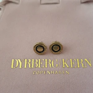 Örhängen Dyrberg/Kern slutsålda  - Guld med svart och text: Travel is to live. ALDRIG ANVÄNDA. 