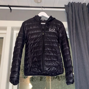 Ea7 Giacca Piumino - BETALA MED SWISH. Giacca Piumino från EA7 är en lättviktsjacka i svart. Med detaljer som huva och en EA7 logga i silver på framsidan. Den är knappt använd 1 säsong och är i topp skick.
